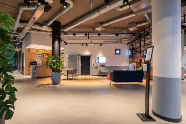 Moderne en open kantoorlobby met een koffiebar genaamd "Zwart Goud", zitgedeelte en zichtbare ventilatiebuizen aan het plafond.